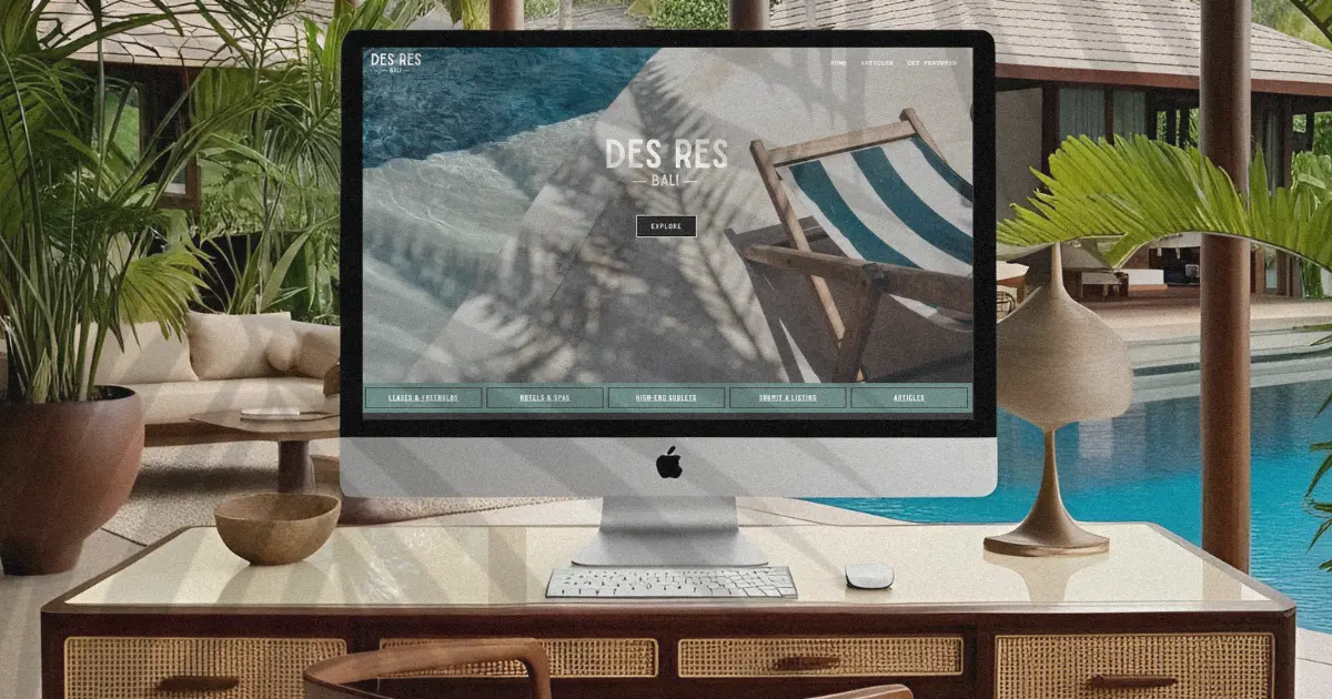 iMac on a wicker-drawer desk inside a Bali villa, showing the Des Res Bali homepage with tropical pool view and palm trees