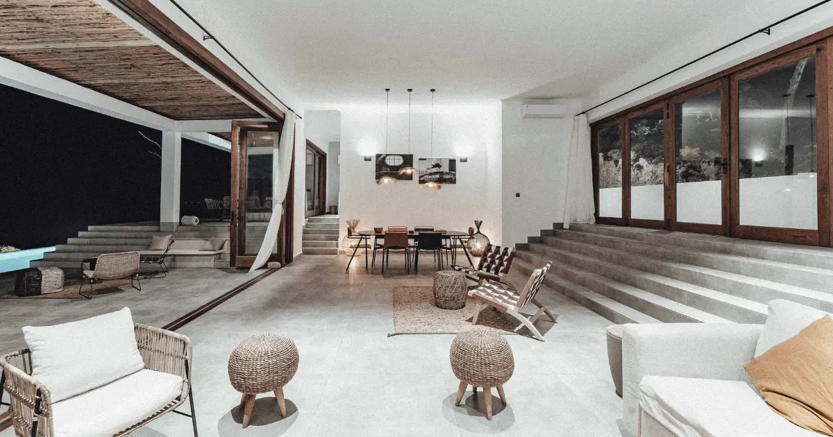 Modern Bali villa interior at night, with concrete walls, bamboo furniture, sunken living room, and a softly lit pool visible through open wood-framed glass doors.