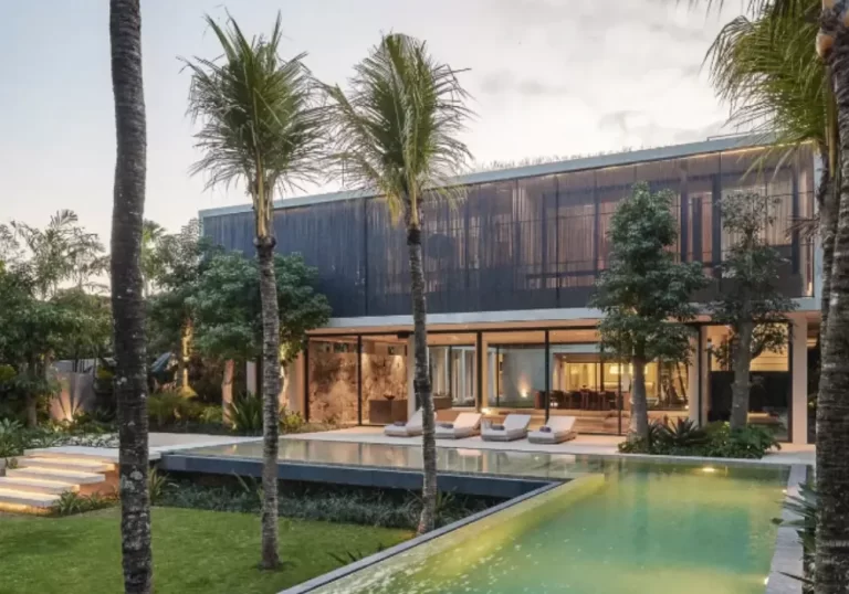 Exterior of a contemporary 6-bedroom villa in Pererenan with dark wood slatted siding, open sliding doors, and a large L-shaped pool lit at night.