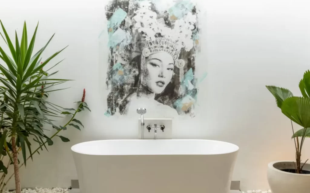 Modern white bathroom at Anwa Bali featuring a freestanding bathtub, tropical palms, and a painting of a Balinese woman in ceremonial headdress.