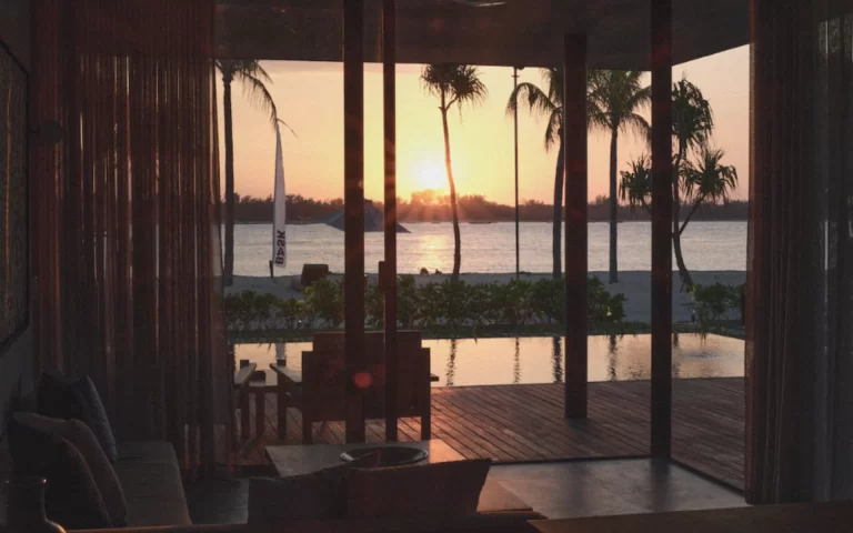 Sunset view from a BASK Gili Meno villa showing a private infinity pool, beachfront deck, and palm trees silhouetted against the horizon