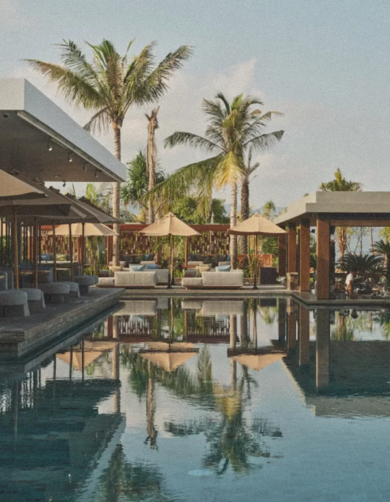 Main swimming pool at BASK Gili Meno with loungers, umbrellas, palm trees, and reflections on a sunny day