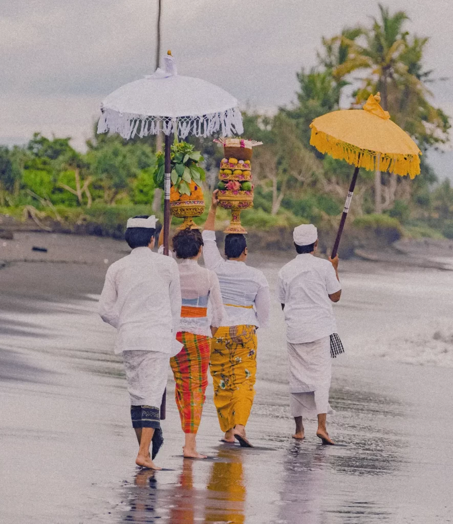 Four Balinese people in ceremonial dress walking on black sand beach carrying traditional offerings and umbrellas