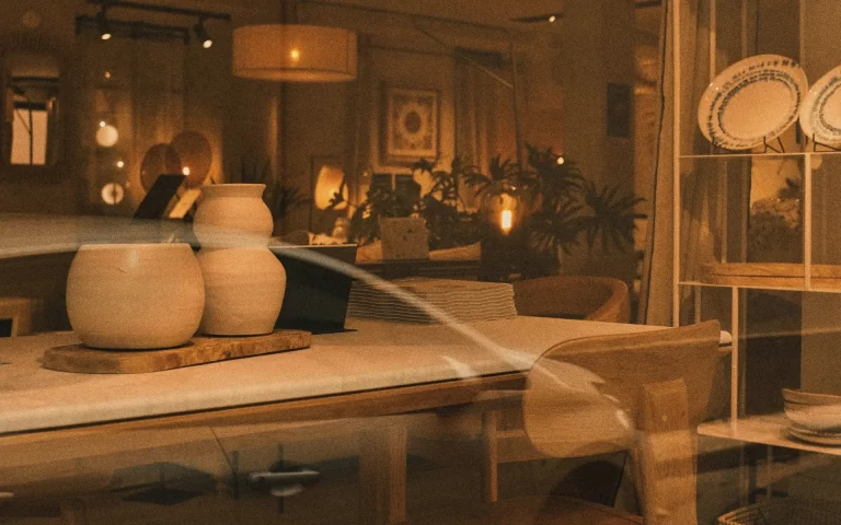 Bali furniture and decor displayed in a shop window with warm golden light and a faint street reflection.