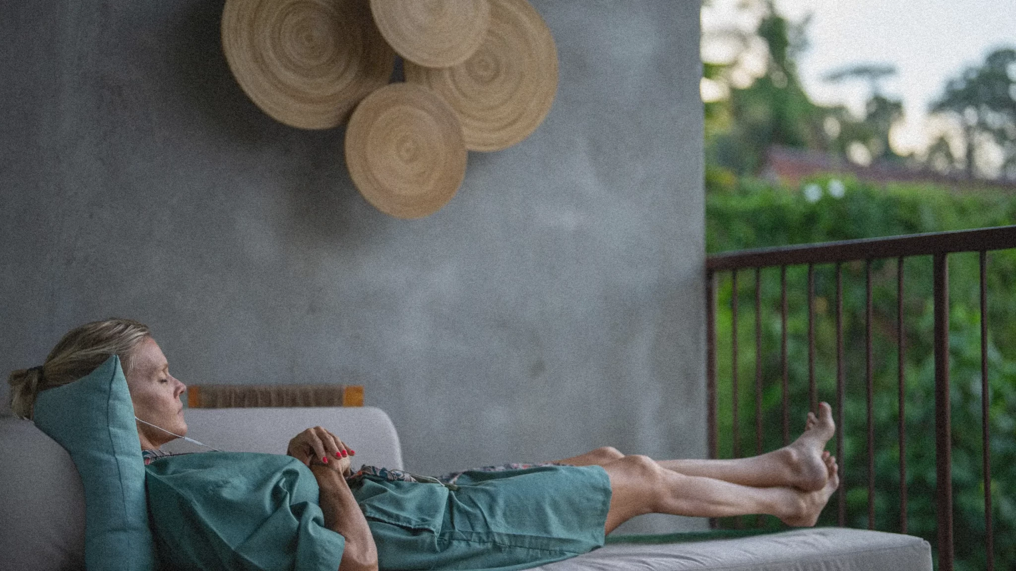 Woman relaxing on Adiwana Hotel balcony lounge, serene with headphones, overlooking lush jungle and palms, minimalist wood wall sculpture in background.