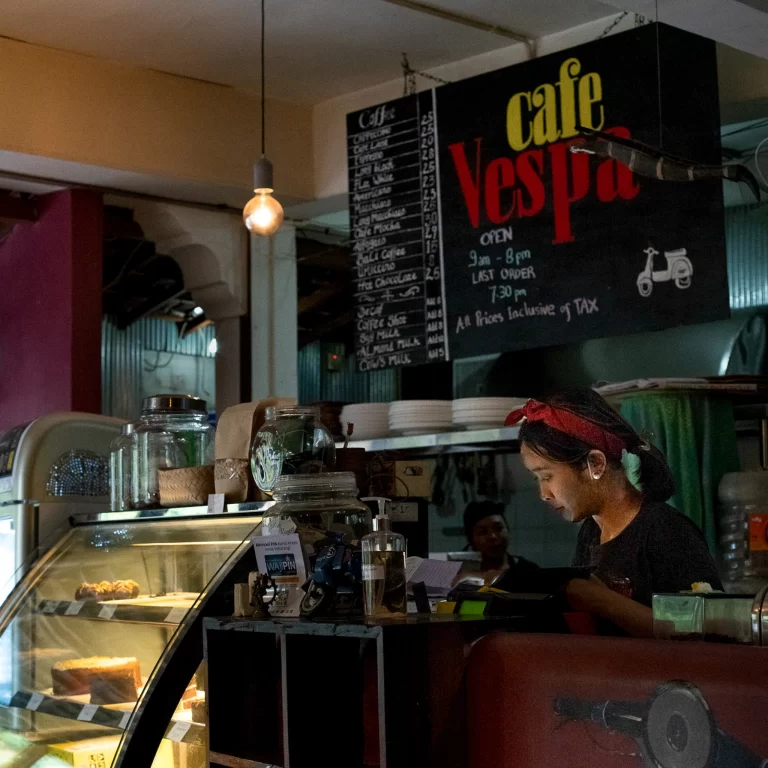 Café Vespa cashier behind counter with illuminated dessert display and coffee menu, cakes visible.