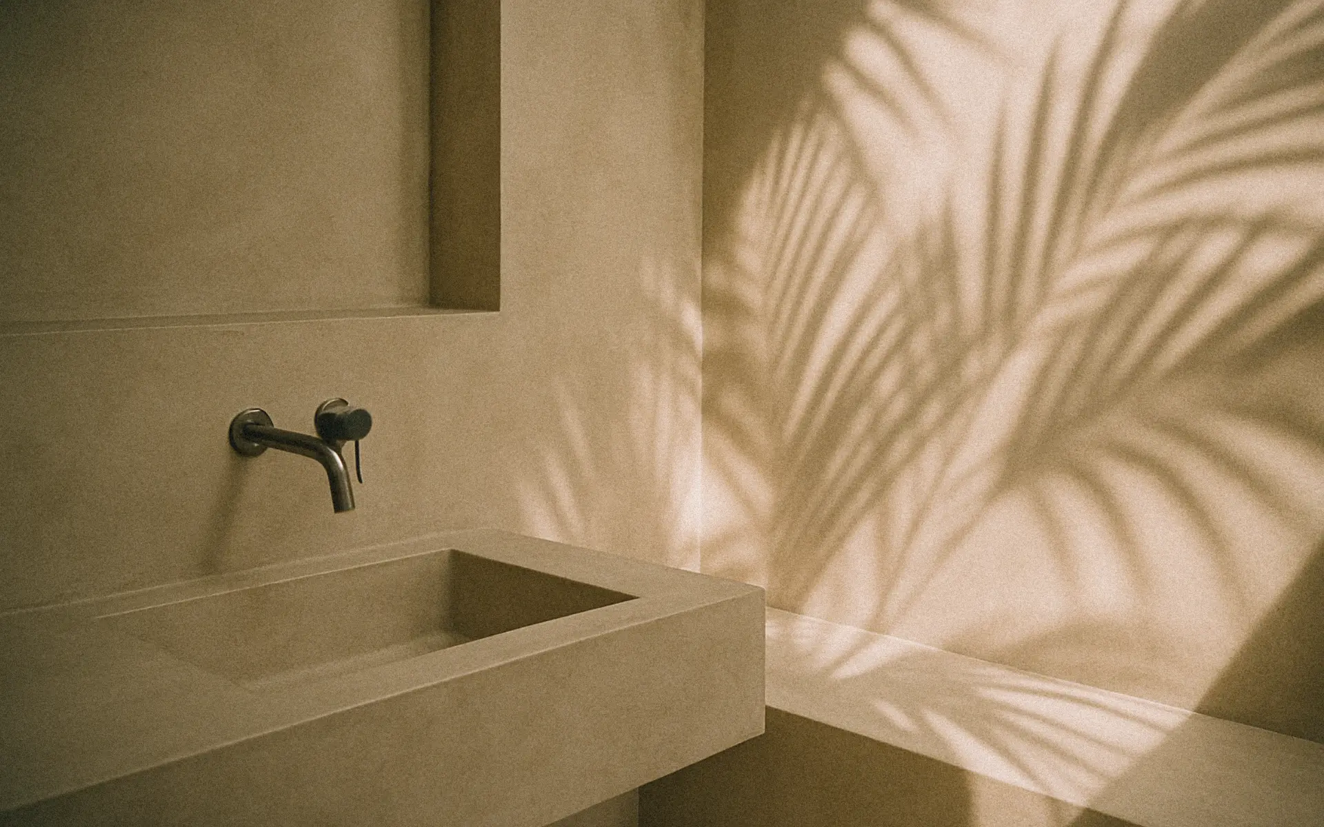 Minimalist concrete bathroom sink with golden light casting palm leaf shadows on the wall.