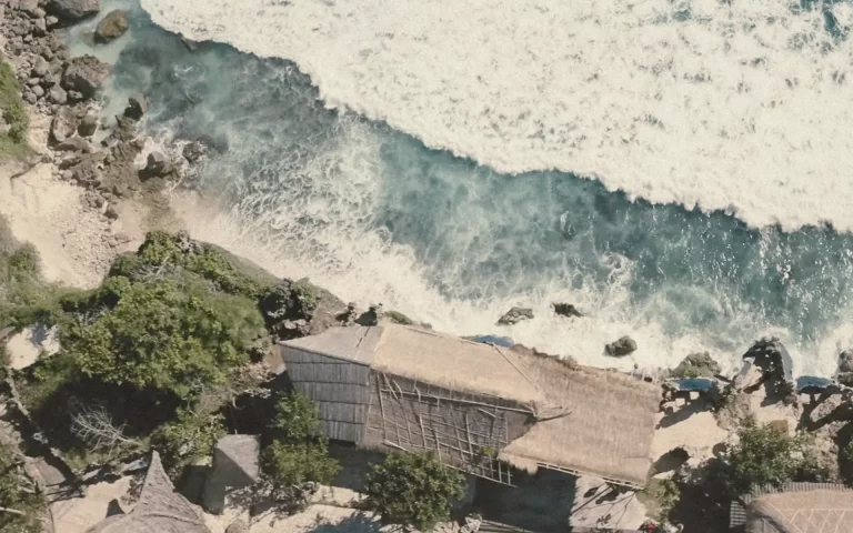 Aerial view of Malini Uluwatu perched atop the cliffs, overlooking the turquoise ocean and jagged coastline, with the building’s terraces and roof visible.
