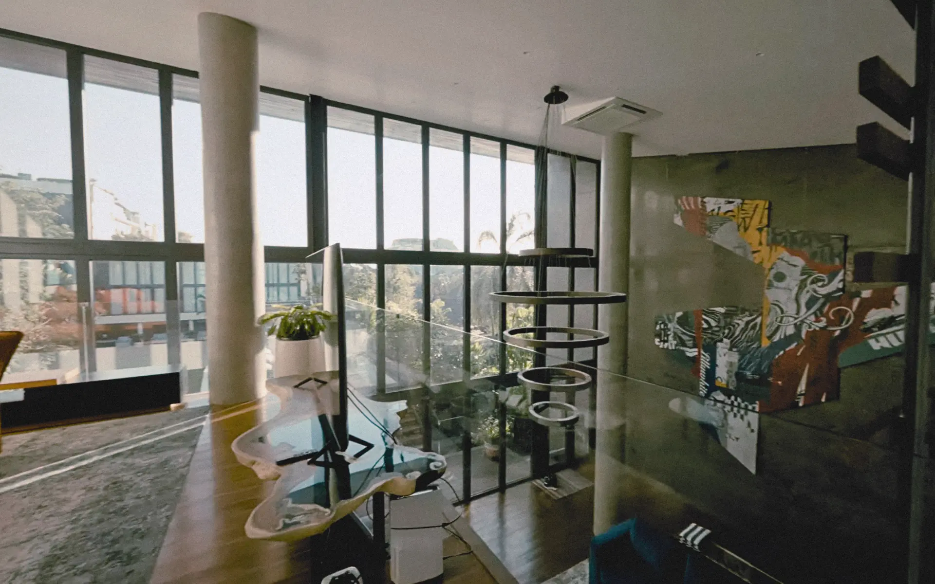 Interior of tropical brutalist villa with exposed concrete walls, ceiling beams, black-paned window, and contemporary Indonesian art installation.