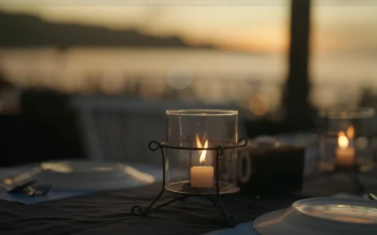 Lit candle on a dinner table at sunset, with blurred plates, people, and Jimbaran beach in the background.