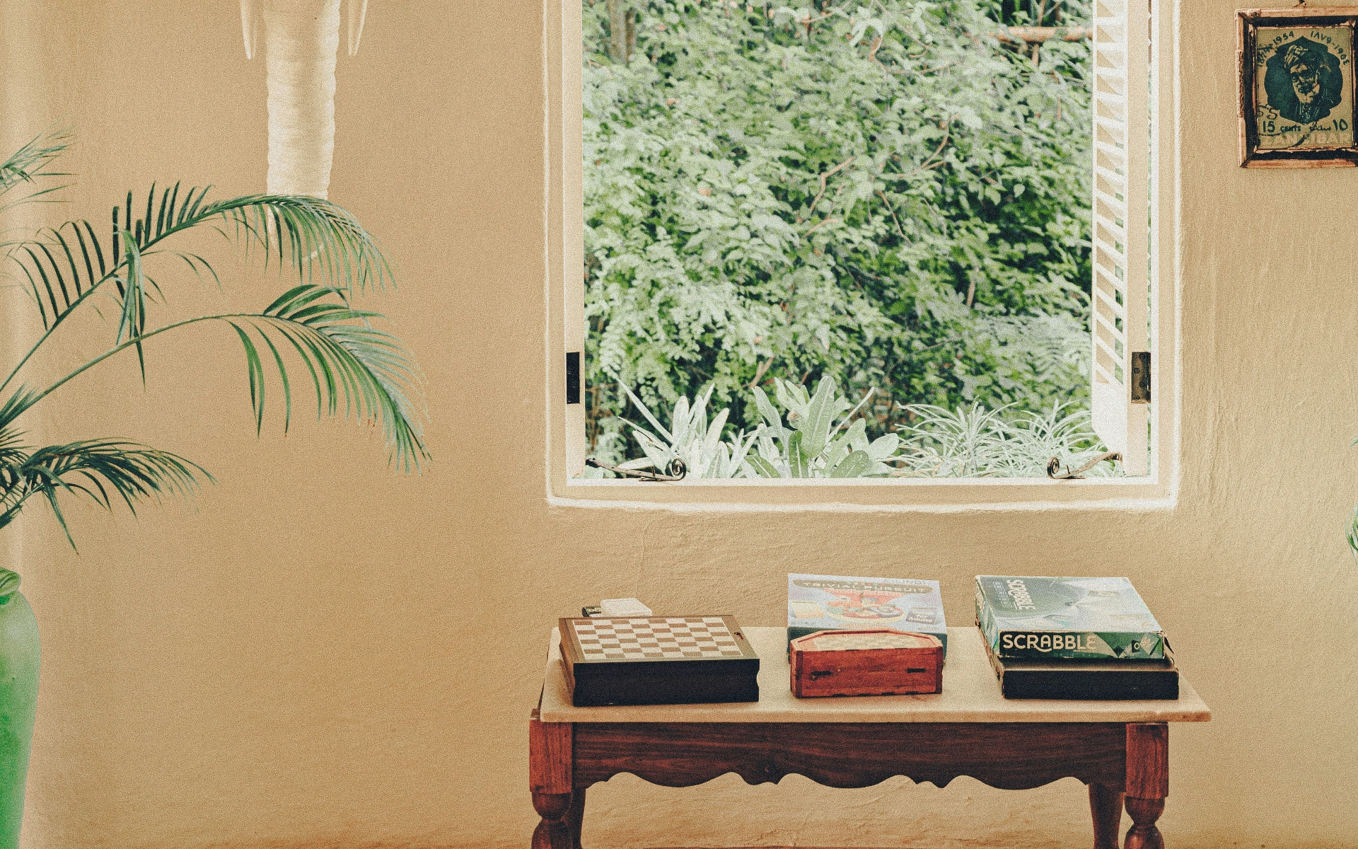 Close-up of a window with open white shutters, indoor palm, vintage table with Scrabble and checkerboard, and framed stamp artwork.