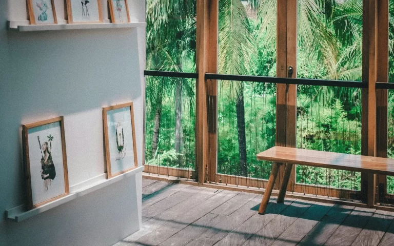 Close-up of framed artwork on wooden wall shelves with wood floors and glass doors opening to a tropical garden.