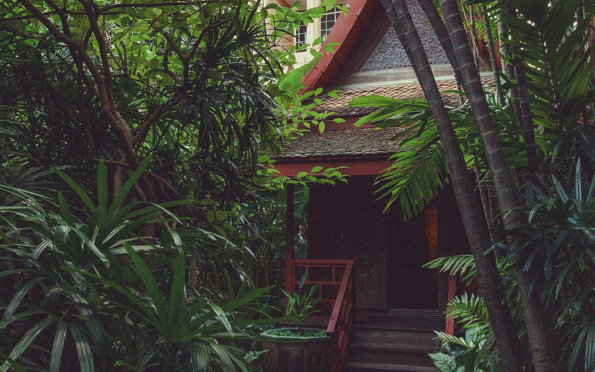 Colonial-style Bali house with red trim and railings, partially hidden behind tropical palms and lush greenery.