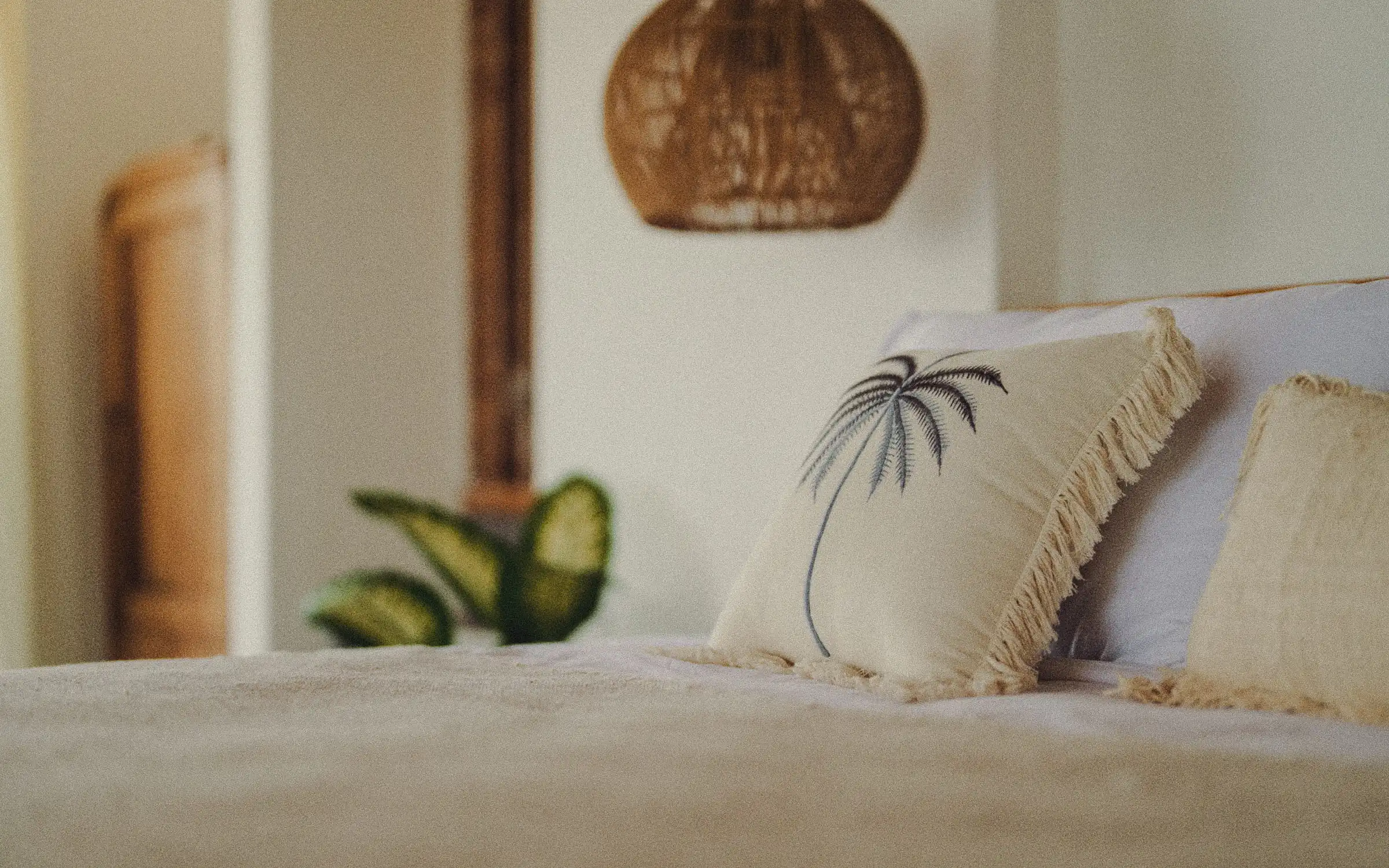 Cream-colored linen bedding with earth-tone sheets, a palm-print cushion, and a rattan pendant lamp in a Bali bedroom