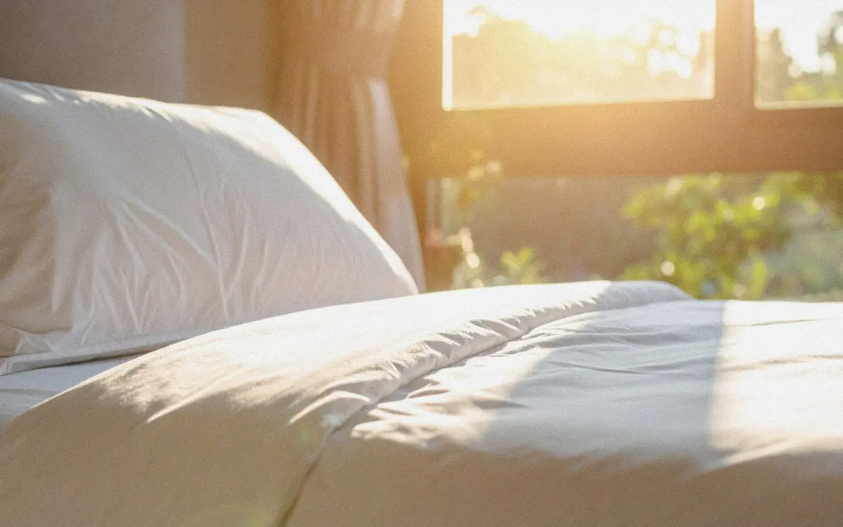 Close-up of a bed with white linen sheets and pillows, soft golden light filtering through a nearby window