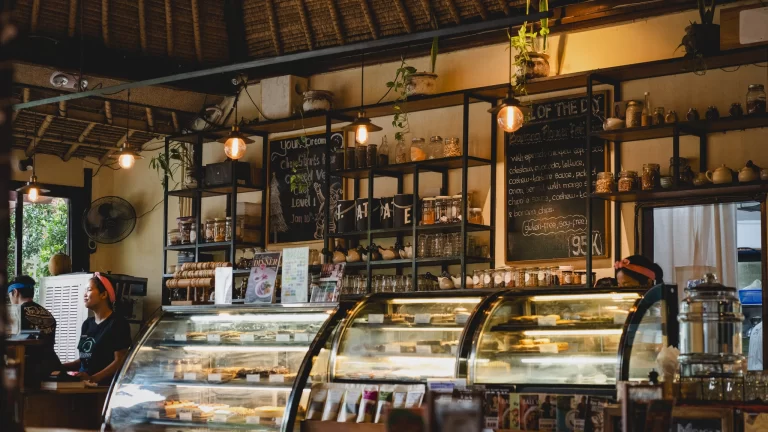 Main counter at Sayuri Ubud with staff behind the glass dessert display, chalkboard specials, hanging plants, shelves of tea and Expat coffee, warm earthy lighting.
