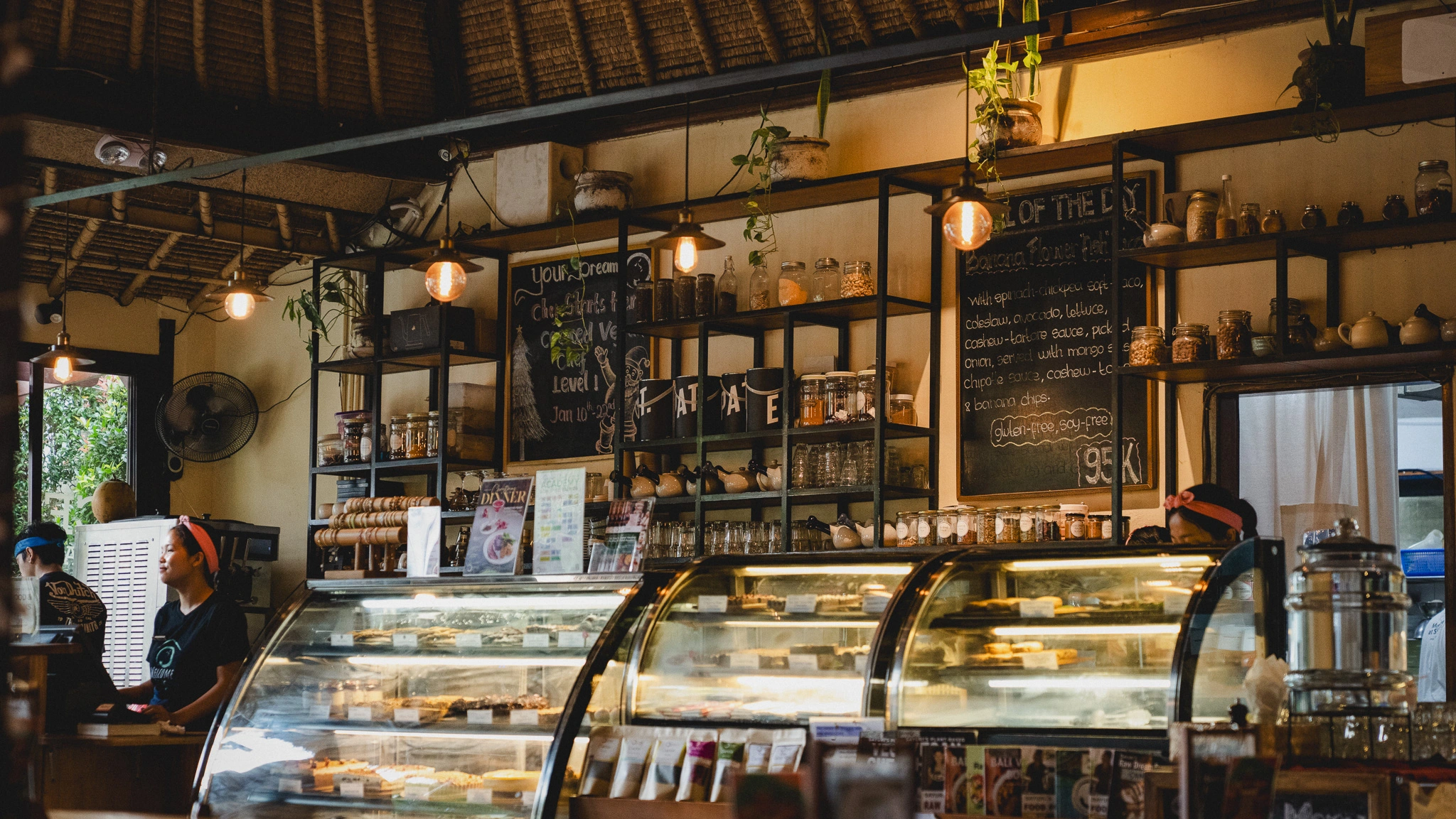 Main counter at Sayuri Ubud with staff behind the glass dessert display, chalkboard specials, hanging plants, shelves of tea and Expat coffee, warm earthy lighting.