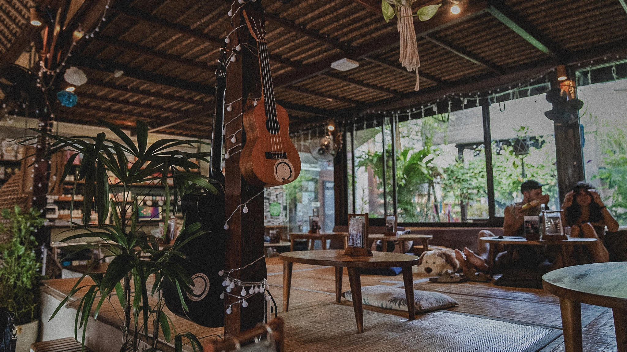 Interior of Sayuri Ubud showing the upper platform seating area, a few guests, pillows on the floor, a guitar on the wall, plants around, sunlight streaming in, relaxed vibe.