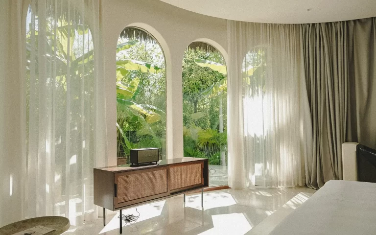 Tropical bedroom with white walls, arched windows, sheer curtains and mid-century wood furniture overlooking lush foliage in Bali