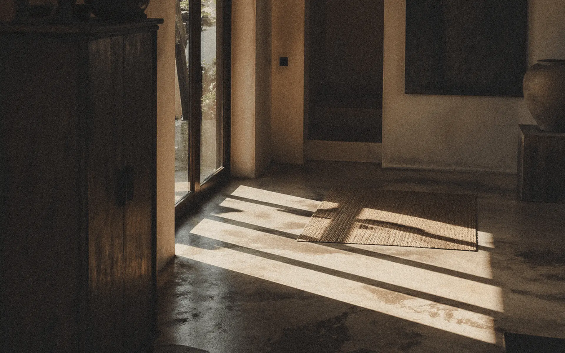 Filtered sunlight through glass doors casting geometric shadows on a terracotta floor inside a Bali villa.