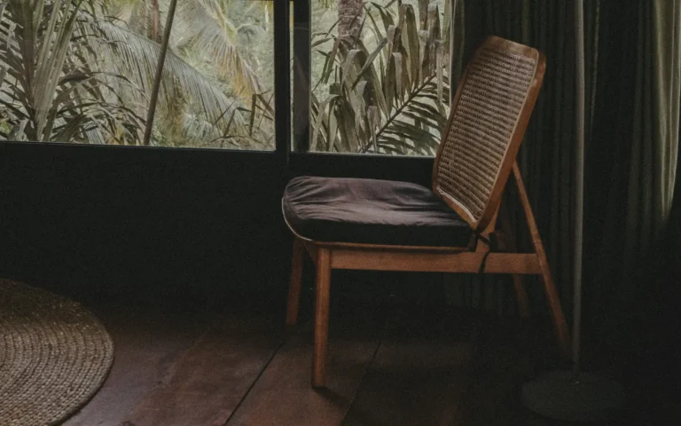 Moody rattan and wood chair beside a tropical window with palm shadows and filtered sunlight