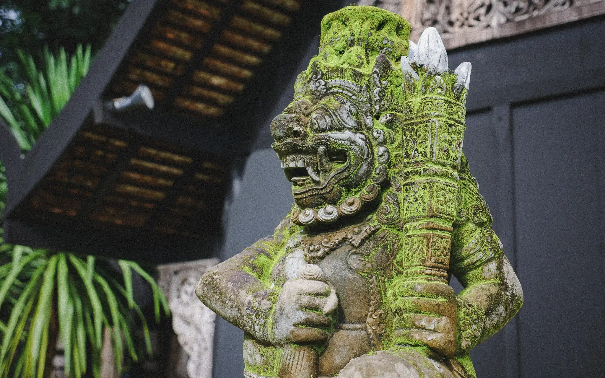Moss-covered Balinese guardian statue surrounded by tropical architecture and lush greenery in Bali, Indonesia.