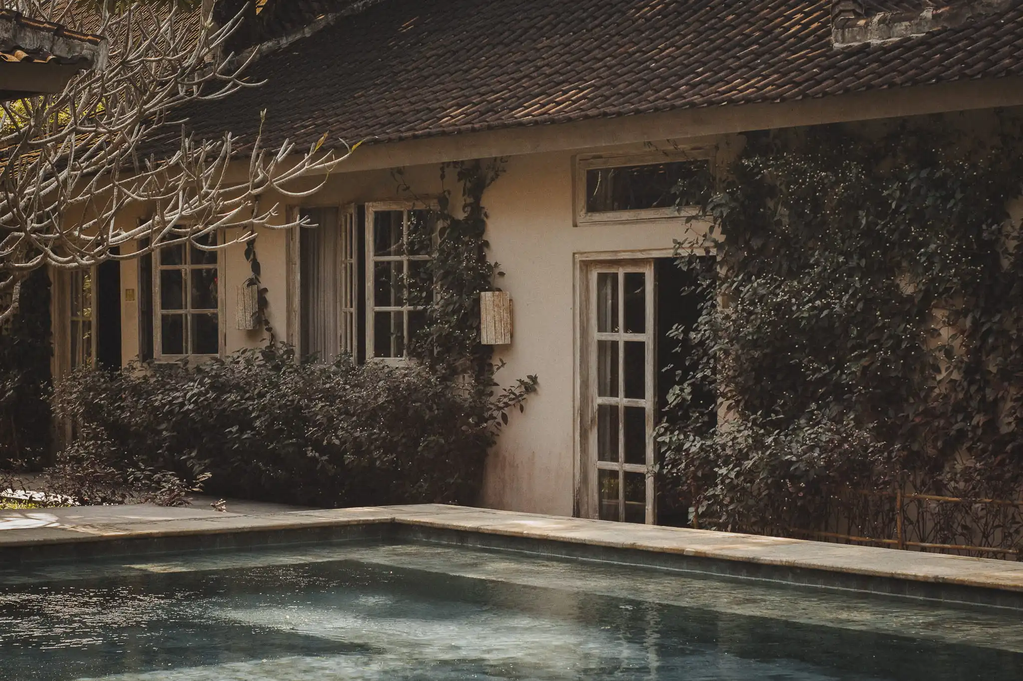 Older house with weathered timber windows, tiled roof and established planting beside a swimming pool