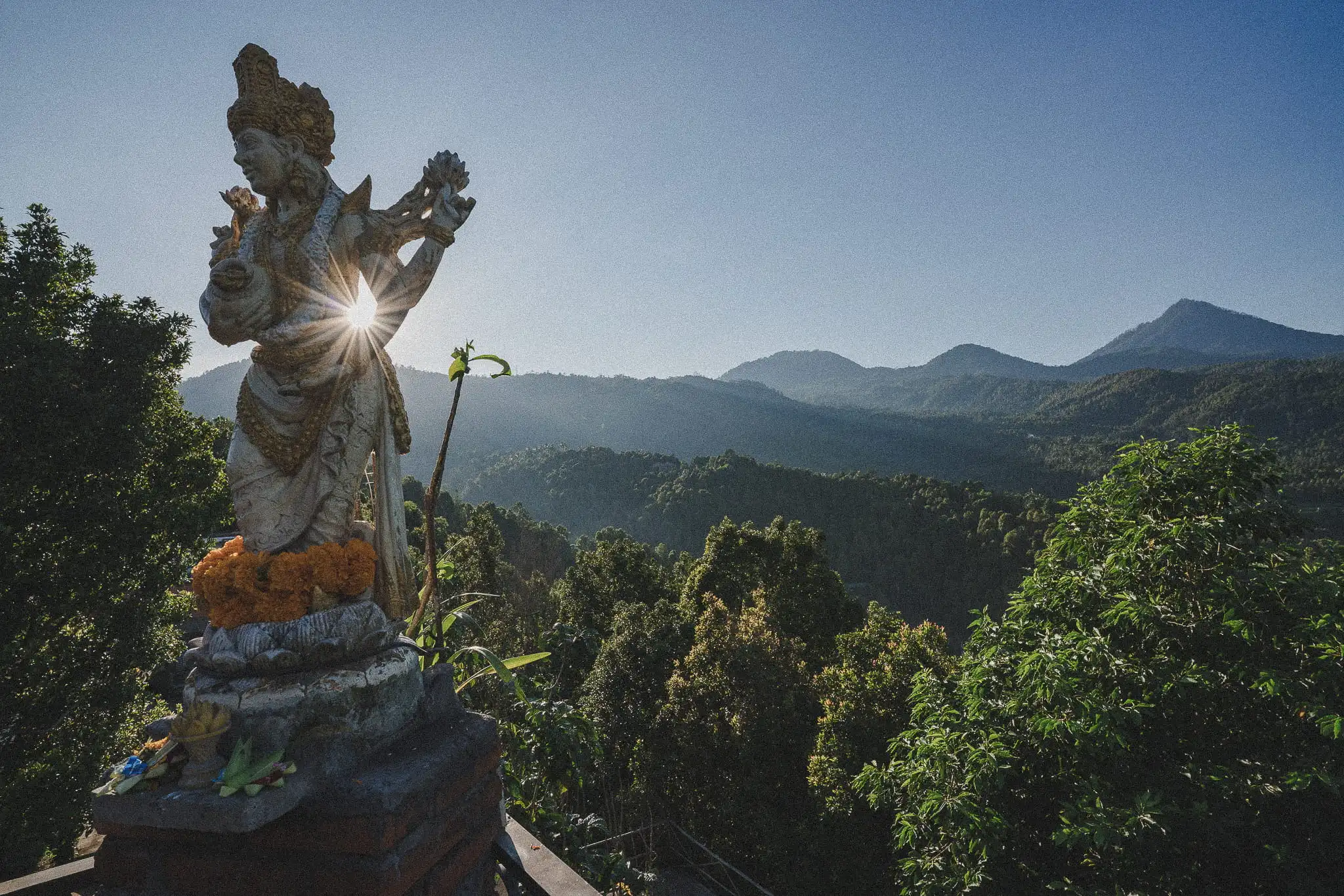 Mountain landscape in Munduk, Bali, with a temple statue silhouetted by morning sunlight